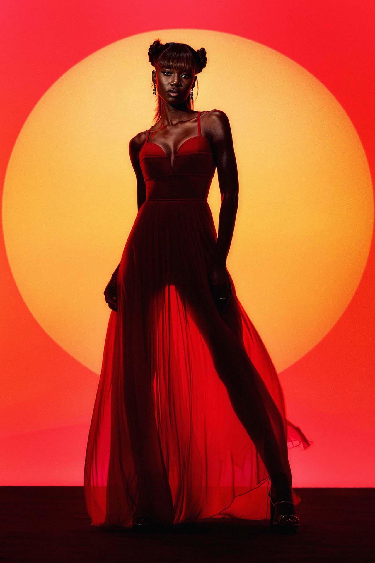A full-body silhouette pictorial of a model in a red sheer dress against a large, circular orange sun-like backdrop.