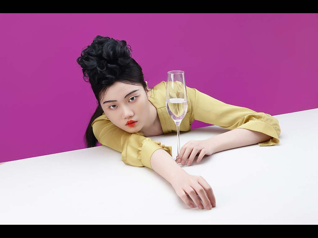 보라색 배경 앞에서 노란색 블라우스를 입은 모델이 샴페인 잔과 함께 테이블에 기대어 포즈를 취하고 있는 감각적인 패션 사진