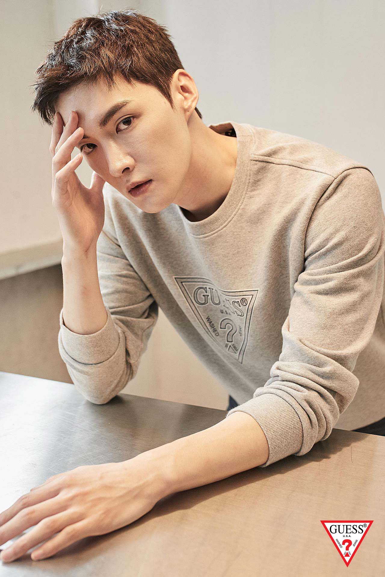 한글: 게스 로고가 새겨진 그레이 스웨트셔츠를 입고 테이블에 기대어 정면을 응시하는 남성 모델의 브랜드 광고 사진. English: A brand advertisement photo of a male model in a gray GUESS logo sweatshirt, leaning on a table and gazing at the camera.