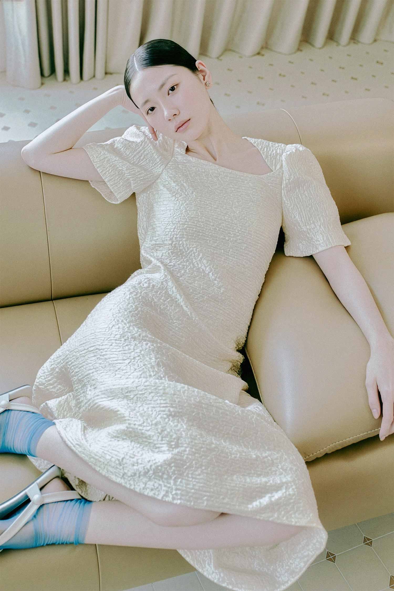 포토그래퍼 김준형이 캡처원 워크플로우로 기획 및 촬영한 여성 패션 룩북 화보로, 크림색 커튼 배경의 베이지색 가죽 소파에 기대어 앉아 셔링 질감의 크림 화이트 스퀘어넥 원피스와 파란색 양말을 착용한 여성 모델의 포트폴리오 사진 (phjun.co.kr)