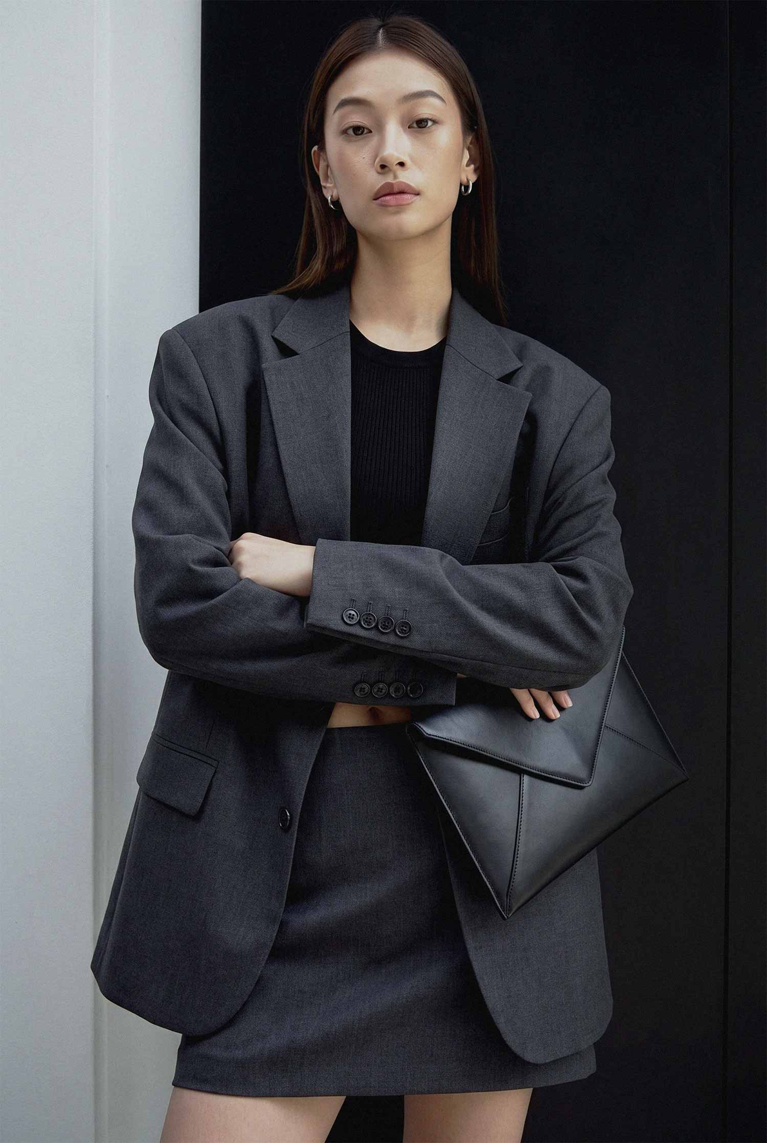 🇰🇷 한국어 Alt Text: 화이트와 블랙 벽면이 분할된 미니멀한 배경 앞에서 오버사이즈 다크 그레이 블레이저 자켓과 미니스커트를 착용하고 블랙 엔벨롭 클러치 백을 든 여성 모델의 패션 룩북 화보. phjun.co.kr 포트폴리오. 🇺🇸 영어 Alt Text: A minimalist fashion lookbook photo of a female model wearing an oversized charcoal grey blazer with a matching mini skirt, holding a black geometric envelope clutch bag against a split black and white background for phjun.co.kr.